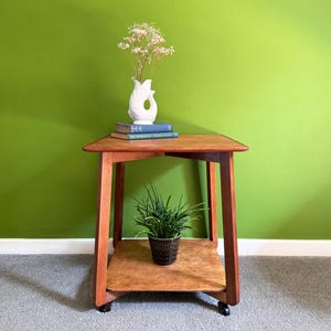 May include: A wooden side table with a lower shelf and a top surface. A white vase with flowers, two blue books, and a potted plant are on the table. The table is brown and has a simple, classic design.
