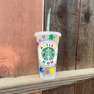 May include: A clear plastic cold cup with a lid and straw. The cup features colorful puzzle pieces, hearts, and the text "AUTISM MOM" in a circular design with a green logo. The cup is decorated with red, yellow, blue, and green accents.