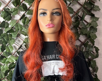 May include: A long, wavy, bright orange wig on a mannequin head. The wig is styled in loose waves and has a natural-looking hairline. The mannequin is wearing a black t-shirt with the text "SA HAIR CREATIONS" printed on it.