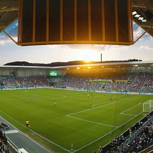 May include: A large crowd of people in a stadium watching a soccer game. The field is green and the players are wearing blue and white uniforms. The sun is setting in the background.