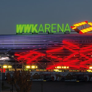Könnte beinhalten: Nachtansicht der WWK ARENA mit grünen, roten und violetten Lichtern. Der Name des Stadions ist in großen grünen Buchstaben zu sehen. Moderne Architektur mit geometrischem Muster.