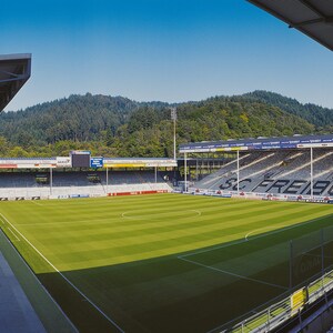 May include: A wide-angle view of a soccer stadium with green grass, white lines, and a large sign that reads "SC FREIBURG". The stadium has a large, covered seating area on the left side of the image and a smaller, uncovered seating area on the right side.