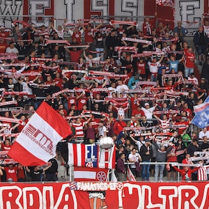 Puede incluir: Un gran grupo de personas con bufandas y camisas rojas y blancas animan en un evento deportivo. La multitud sostiene banderas y pancartas. La pancarta en primer plano dice "EINFACH DIE ULTRAS 1991".