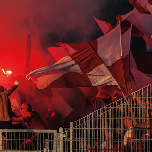Puede incluir: Una multitud de personas en un estadio, sosteniendo bengalas rojas y ondeando banderas blancas y rojas. La escena está llena de humo y emoción.