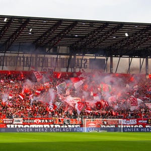 May include: A stadium filled with a crowd of people, many waving red and white flags. Smoke and flares add to the atmosphere. The stands are packed, and the field is green.