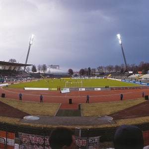 Puede incluir: Una vista panorámica de un estadio de fútbol con un campo verde, una pista roja y una gran multitud de espectadores. El estadio tiene una gran estructura de techo blanco y dos postes de luz altos.