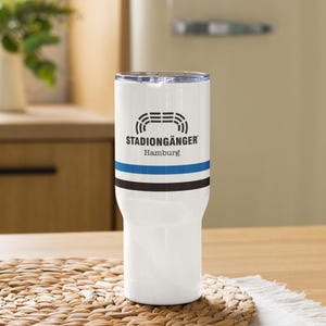 May include: White insulated tumbler with a black and blue stripe design. The tumbler features the text "STADIONGÄNGER Hamburg" in black. The tumbler is on a wooden surface.
