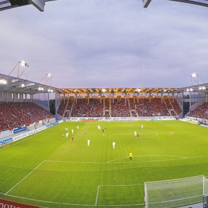 Könnte beinhalten: Eine Weitwinkelaufnahme eines vollen Fußballstadions mit einem grünen Rasen und einem weißen Tor im Vordergrund. Die Tribünen sind mit Fans gefüllt, die rot und weiß tragen.