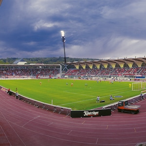 Puede incluir: Una vista de gran angular de un estadio de fútbol con una pista de atletismo roja y un campo verde. Las gradas están llenas de espectadores y hay varios jugadores en el campo. El estadio está iluminado por focos.