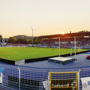 May include: A large outdoor stadium with a green grass field and a blue running track. The stadium is filled with people watching a game. The sun is setting in the background.
