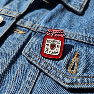 May include: A red enamel pin shaped like a jar of jam with a pink lid. The label on the jar is white with a red strawberry graphic, two small red hearts, and the black text READING IS MY JAM. The pin is attached to a blue denim fabric surface.