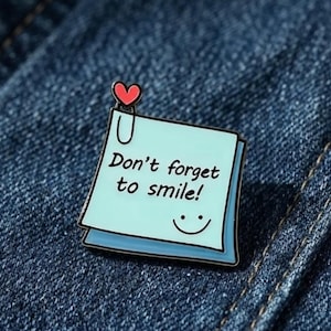 May include: A light blue square enamel pin shaped like a sticky note with a black outline. The pin features the text "Don't forget to smile!" in black script, a small black smiley face, and a red heart attached to a black paperclip at the top.