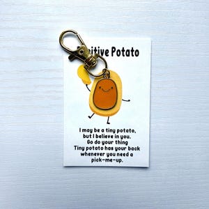 May include: A gold-coloured keychain featuring a cartoon potato charm and a card. The card reads "Positive Potato" and includes an inspirational message. The potato charm is yellow and brown with a smiling face.