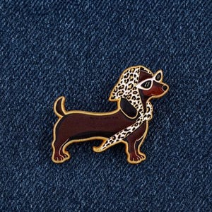 May include: A brown dachshund enamel pin featuring a leopard print headscarf and white-rimmed sunglasses. The dog is depicted in profile with gold-toned metal outlines, showcasing a sleek body, short legs, and a curled tail against a blue denim background.