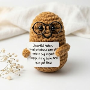 May include: A handmade crochet potato toy, wearing round black glasses and a smiling expression. It holds a sign with the text: "Cheerful Potato, Small potatoes can still make a big impact! Keep pushing forward, you got this!"