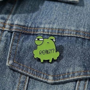 May include: A green enamel pin shaped like a frog with a sceptical expression, featuring large eyes and the word "REALLY?" printed in black text on its body. The pin is attached to a blue denim fabric surface with visible stitching and texture.