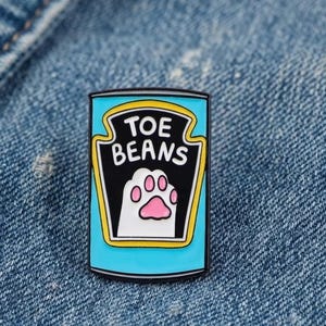 May include: A rectangular enamel pin featuring a light blue can design with a black label outlined in yellow. The text TOE BEANS is printed in white above a white cat paw illustration with pink pads, mimicking a classic food can label aesthetic.
