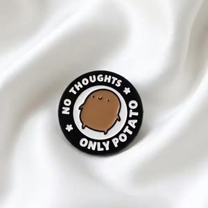 May include: A round black enamel pin featuring a central brown cartoon potato character with a simple smiling face. The text NO THOUGHTS ONLY POTATO is printed in white capital letters around the border, accented by two small white stars.