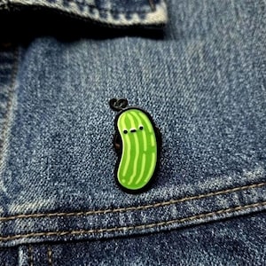 May include: A small enamel pin shaped like a green pickle with a black outline and a simple face consisting of two black dots for eyes and a small horizontal line for a mouth, attached to a blue denim fabric surface with visible stitching.
