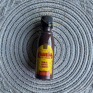 May include: A small bottle of Kahlúa rum and coffee liqueur. The bottle has a black cap and a gold label with the brand name and product information. The bottle is filled with a dark brown liquid and is decorated with a gold string.