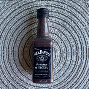 May include: A miniature Jack Daniel's whiskey bottle pendant. The bottle is filled with a dark liquid and has a black cap. The label reads "Jack Daniel's Old No. 7 Brand Tennessee Sour Mash Whiskey". The pendant hangs from a silver chain.