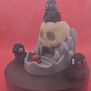 May include: A 3D-printed fantasy scene featuring a skull, ravens, and a dice. The skull is beige, with a gray mountain-like structure. Three black ravens are positioned around the skull. A red dice is inside the skull's mouth. The base is dark gray.