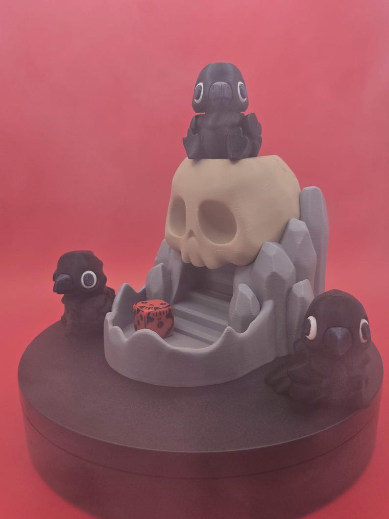 May include: A 3D-printed fantasy scene featuring a skull, ravens, and a dice. The skull is beige, with a gray mountain-like structure. Three black ravens are positioned around the skull. A red dice is inside the skull's mouth. The base is dark gray.