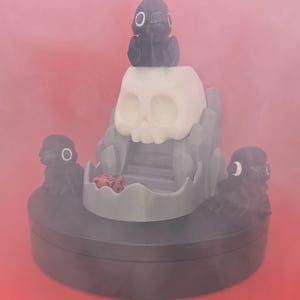 May include: A gray and white skull-shaped sculpture with a small black figure on top and two on the sides. A red and black die is inside the skull's mouth. The sculpture sits on a dark gray base against a red background.