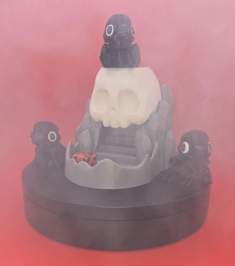 May include: A gray and white skull-shaped sculpture with a small black figure on top and two on the sides. A red and black die is inside the skull's mouth. The sculpture sits on a dark gray base against a red background.
