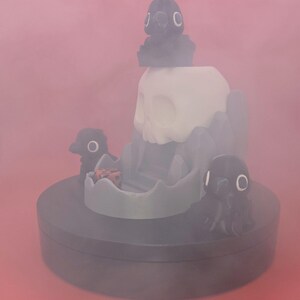 May include: A whimsical, dark gray dice tower with a skull and crow design. The tower has a white skull, gray rock formations, and three crow figures. A red and black die sits in the tower's tray. The base is dark gray.