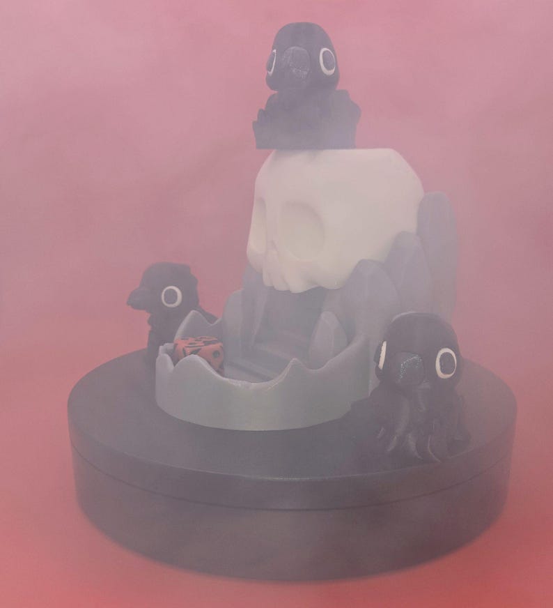 May include: A whimsical, dark gray dice tower with a skull and crow design. The tower has a white skull, gray rock formations, and three crow figures. A red and black die sits in the tower's tray. The base is dark gray.
