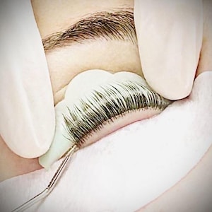 Puede incluir: Primer plano de un ojo con un protector de pestañas blanco en su lugar. El protector se utiliza para proteger las pestañas inferiores durante la aplicación de extensiones de pestañas.