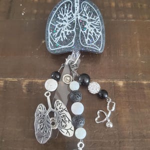 May include: Black and white resin badge reel with a white outline of lungs, black lava rock beads, white beads, a silver charm of lungs, a silver stethoscope charm, and a silver charm with a caduceus and the letters RRT.