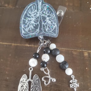 May include: A black and white glitter resin badge reel with a white outline of lungs. The reel has a black and white beaded chain with a stethoscope charm, a heart charm, and a silver charm with the letters "RN" and a caduceus.