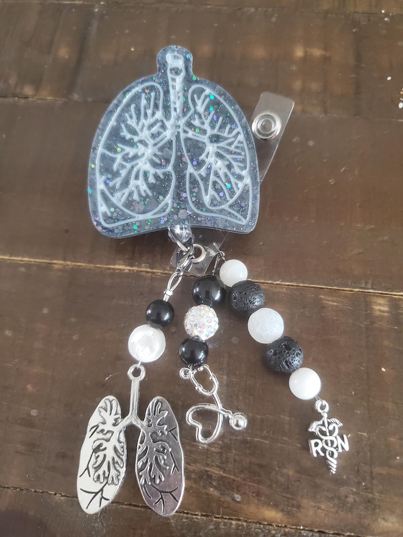 May include: A black and white glitter resin badge reel with a white outline of lungs. The reel has a black and white beaded chain with a stethoscope charm, a heart charm, and a silver charm with the letters "RN" and a caduceus.
