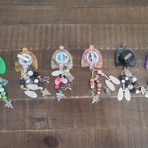 May include: Six colorful retractable badge reels with various charms and beads. The reels are made of clear resin with different colored glitter and designs. The charms include hearts, flowers, and medical symbols. The beads are made of glass, metal, and plastic.