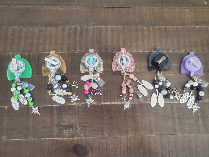 May include: Six colorful retractable badge reels with various charms and beads. The reels are made of clear resin with different colored glitter and designs. The charms include hearts, flowers, and medical symbols. The beads are made of glass, metal, and plastic.