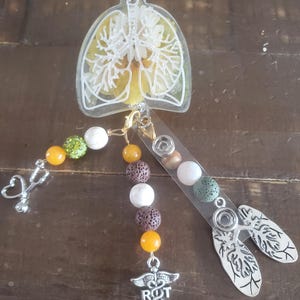 May include: A clear resin charm with a white outline of lungs, filled with yellow and white dried flowers. The charm is attached to a beaded lanyard with orange, brown, white, and green beads. The lanyard also has a silver charm with a medical symbol and the letters "R.T."