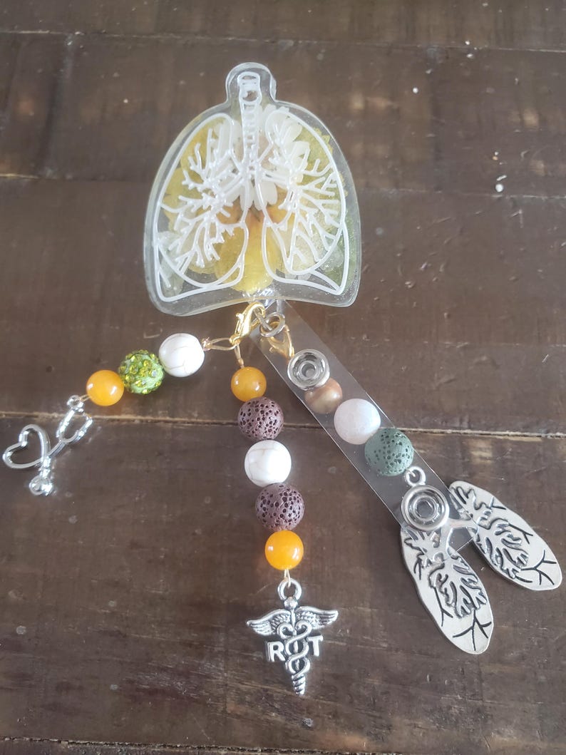 May include: A clear resin charm with a white outline of lungs, filled with yellow and white dried flowers. The charm is attached to a beaded lanyard with orange, brown, white, and green beads. The lanyard also has a silver charm with a medical symbol and the letters "R.T."