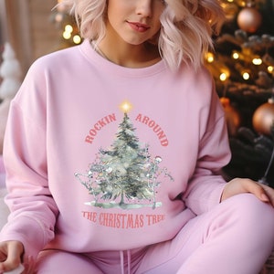 May include: A light pink sweatshirt and matching sweatpants set. The sweatshirt features a Christmas tree design with the text "ROCKIN AROUND THE CHRISTMAS TREE". The design includes skeletons and ornaments. The woman in the image is wearing the set.