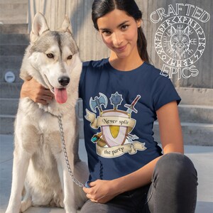 Peut inclure: T-shirt bleu marine avec un motif d'écu orné de haches croisées, d'une masse et d'une épée. La bannière indique "Never split the party". Un chien husky est également présent.