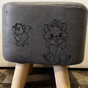 Puede incluir: Un reposapiés gris tapizado con patas de madera. La parte superior del reposapiés presenta un diseño bordado de dos gatos de dibujos animados.