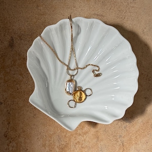 May include: A white, shell-shaped dish contains two gold necklaces and two rings. One necklace features a rectangular pendant with a white stone, and the other has a round, gold pendant. The rings are small and gold.