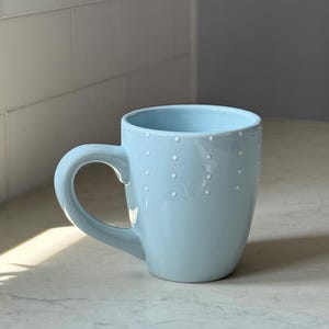 May include: A set of three ceramic mugs with a speckled, matte finish. The mugs are in shades of cream, light gray, and dark gray. Each mug has a simple, minimalist design, perfect for coffee or tea.
