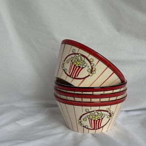 May include: A stack of four ceramic popcorn bowls. Each bowl has a cream-colored background with red vertical stripes. A popcorn graphic is centered on each bowl, with a red and white striped popcorn container and yellow popcorn.