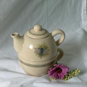 May include: A cream-coloured ceramic tea-for-one set consisting of a teapot stacked on a teacup. The set features a dragonfly illustration, a green leaf-patterned border, and a single pink flower with small yellow blossoms placed at the base.