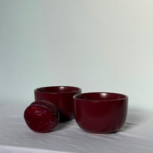 May include: Two deep burgundy ceramic bowls and a ripe plum sit on a white surface. The bowls are round with a smooth finish. The plum is a deep reddish-purple colour.