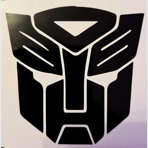 Könnte beinhalten: Schwarz-weiße Grafik des Autobots-Symbols. Das Symbol ist ein stilisierter Robotergesicht mit geometrischen Formen und Linien. Das Design ist kühn und modern, mit einem glänzenden Finish.