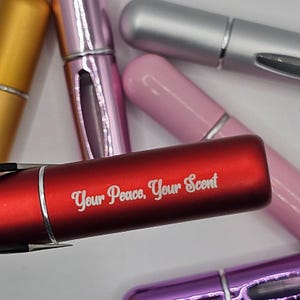 May include: A collection of refillable perfume atomizers in various colors, including red, gold, pink, silver, and purple. The red atomizer features the text "Your Peace, Your Scent" in white lettering. The atomizers have a sleek, cylindrical design.