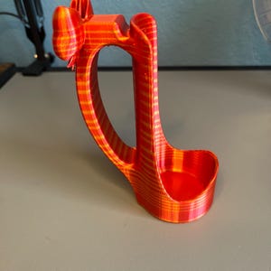 Puede incluir: Un soporte para tel&eacute;fono impreso en 3D, a rayas rojas y amarillas. El soporte tiene un asa curva y una base peque&ntilde;a y redondeada para sostener un tel&eacute;fono. La parte superior tiene un dise&ntilde;o en forma de lazo. El soporte es de pl&aacute;stico.